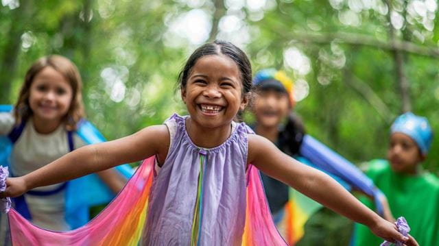 In the forefront, a smiling girl is smiling wearing a cape and pretending to be a dragonfly